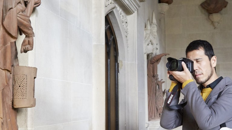 A visitor with a camera up to his face, one eye closed, as he takes a photo of a nearby statue of a figure with a pointy hat and cloak on